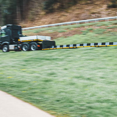 Forsttiefbett Sattelauflieger, der auf einem grasbewachsenen Straßenrand fährt und für den Transport von großen und schweren Forstmaschinen bestimmt ist.